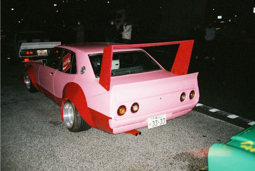 A custom pink Japanese car with a rear spoiler, visible Coca-Cola logo, and license plate number 33-33 at night.