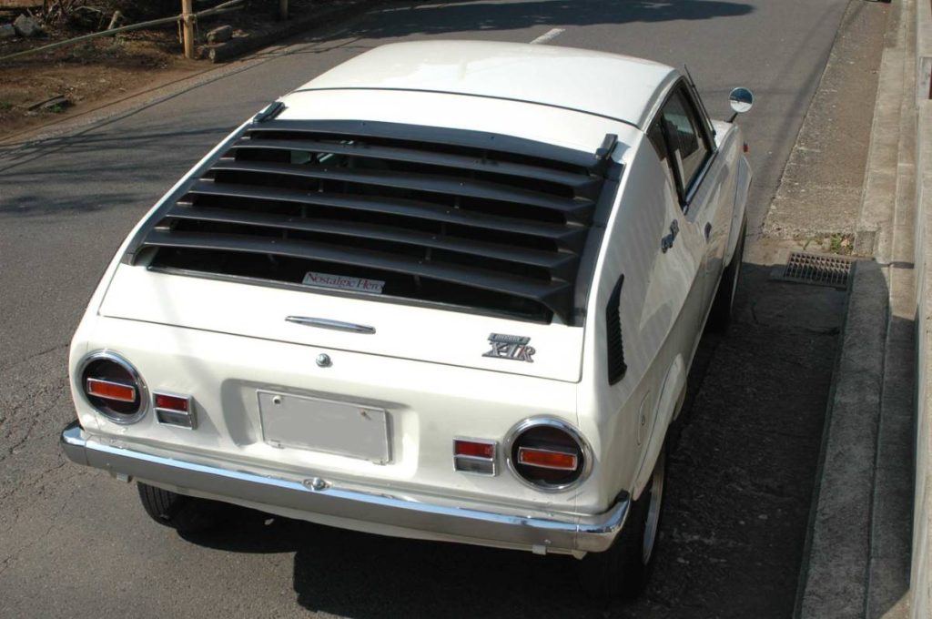 Rear view of a classic white Mazda RX-3 with rear window louvers parked by the road. Text reads Nostalgic Hero.