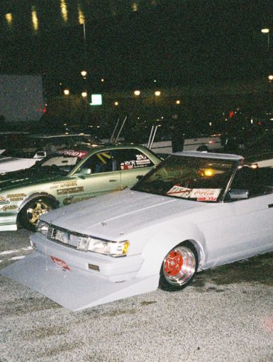 Modified Japanese drift cars parked at night with stickers displaying brand names like WORK and Coca-Cola on the windshields.