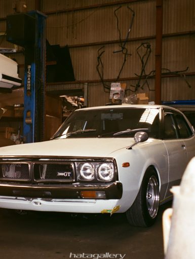 A classic white 2000GT car in a garage with another white car on a lift in the background, hatagallery watermark.