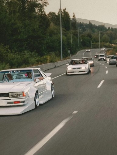 A convoy of modified JDM cars with R.B.S decals on a highway, surrounded by lush greenery, showcasing car culture.
