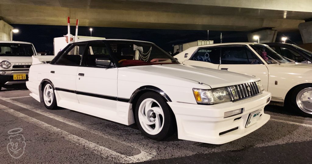 A white classic Japanese car parked under a bridge at night, showcasing its custom body kit and retro design.