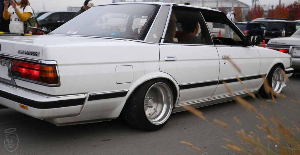 White Toyota Mark II with modified wheels at a car meet, showcasing classic Japanese automotive culture.