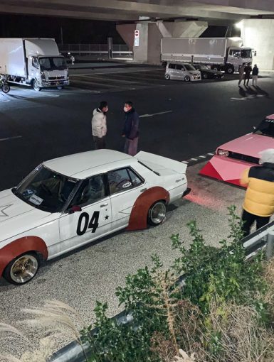 Vintage Japanese modified cars parked under an overpass at night with people standing nearby.