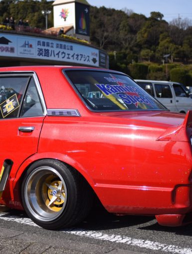 Red customized car with yellow pipes, gold rims, Kaminari Special rear window decal, and Japanese signage in the background.