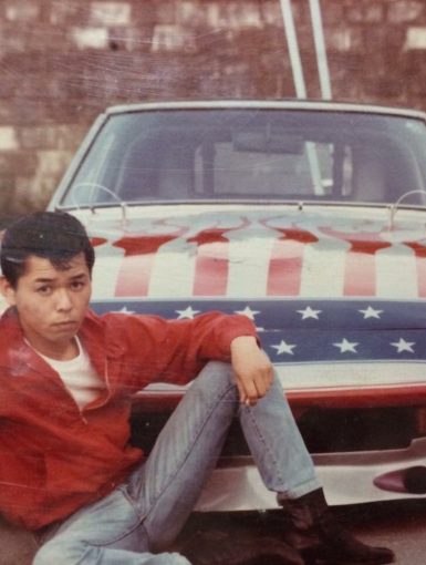 Young man in red jacket poses in front of an American flag-themed car with red flames, white stars, and a blue background.