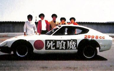 A white sports car with Shinigami and 2998cc written on it, and six people posing behind it on a sunny day.