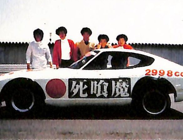 A white sports car with Shinigami and 2998cc written on it, and six people posing behind it on a sunny day.