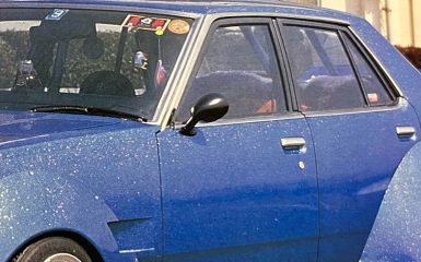 Vintage blue car with Japanese stickers on windshield, showcasing classic design and detailed exterior, captured in daylight.