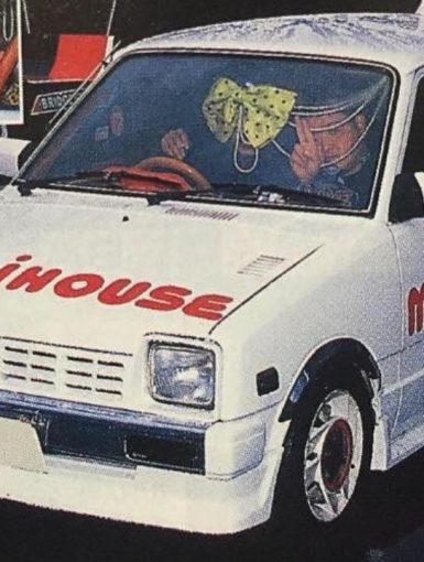 White car with Mikihouse branding, custom rims, bow on windshield, and people inside at an auto show.