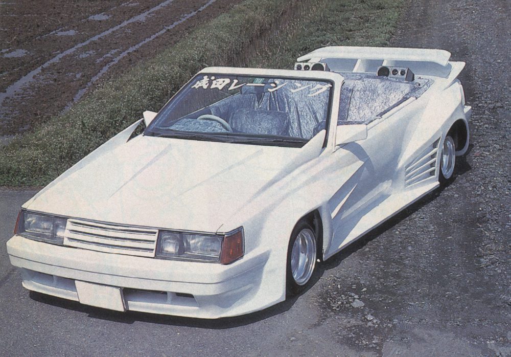 A modified white convertible car with custom body kit and the Japanese text 成田レンジャー on windshield, parked on a rural road.