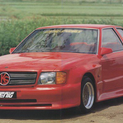 Red Koenig Specials car with sporty design and wide body kit, parked outdoor near a green field.