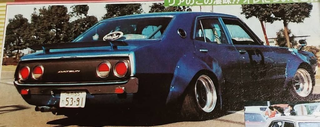 Blue vintage Nissan Skyline Yonmeri car with wide fenders and lowered stance, parked on street with trees in background.