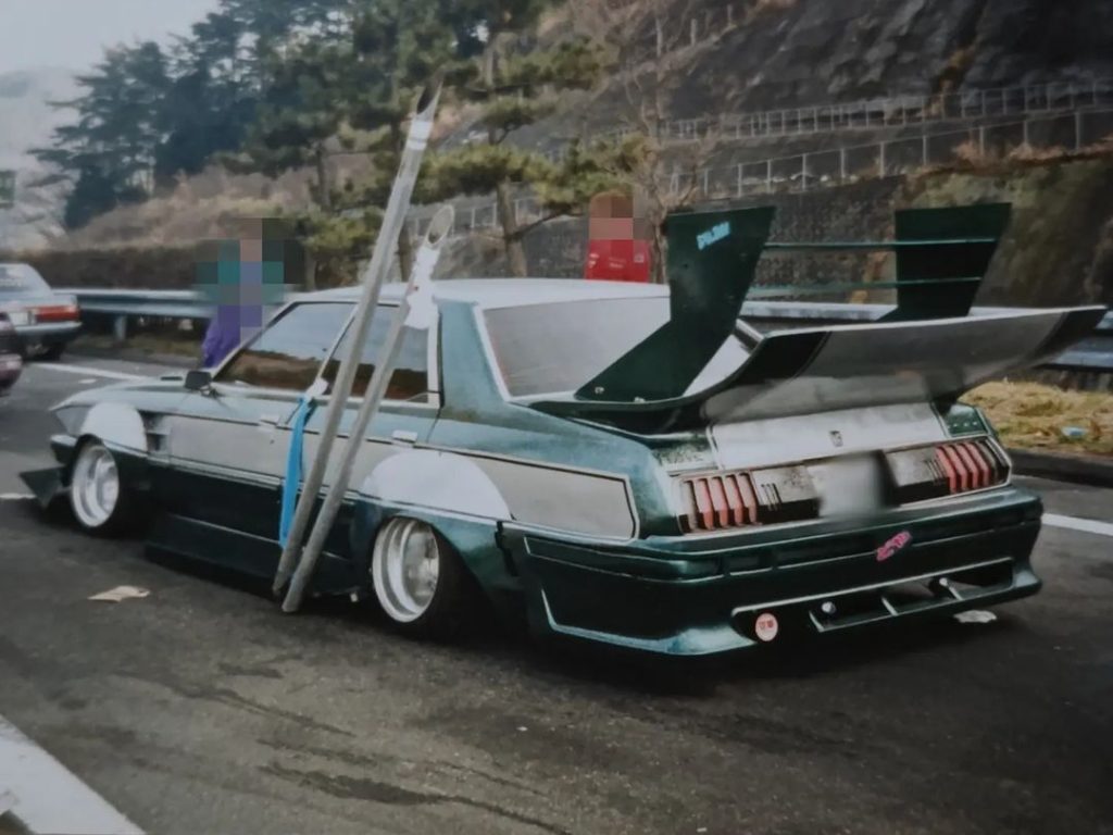 Customized car with a large rear wing and unique modifications stopped on a road with trees and mountains in the background.