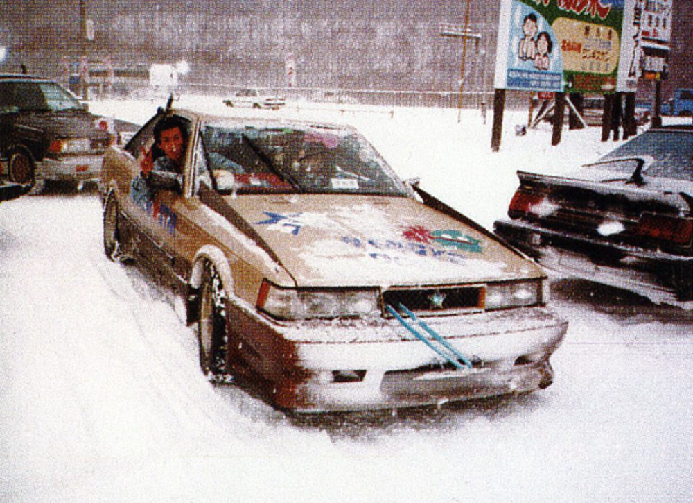 Retro car in snowy landscape, driver leaning out window, snowy road in city with billboards and parked cars.