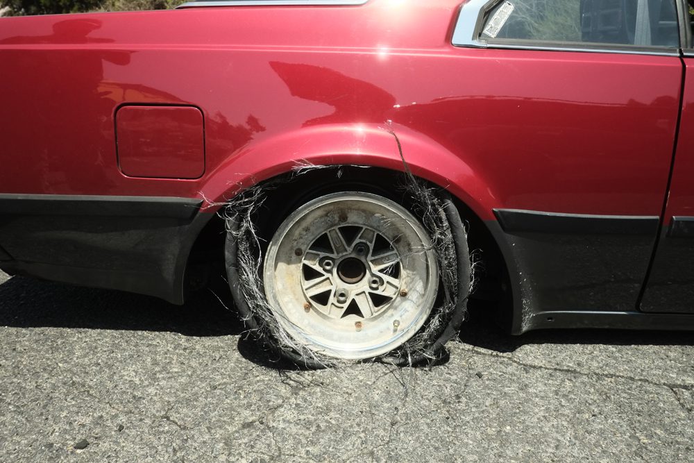 Red car with a severely damaged flat tire on a sunny day, parked on asphalt.