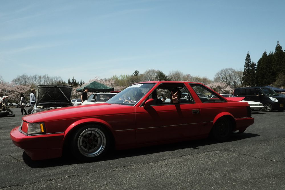 Red Toyota Soarer parked at an outdoor nonsuri meet, surrounded by people and cherry blossom trees under a clear sky.