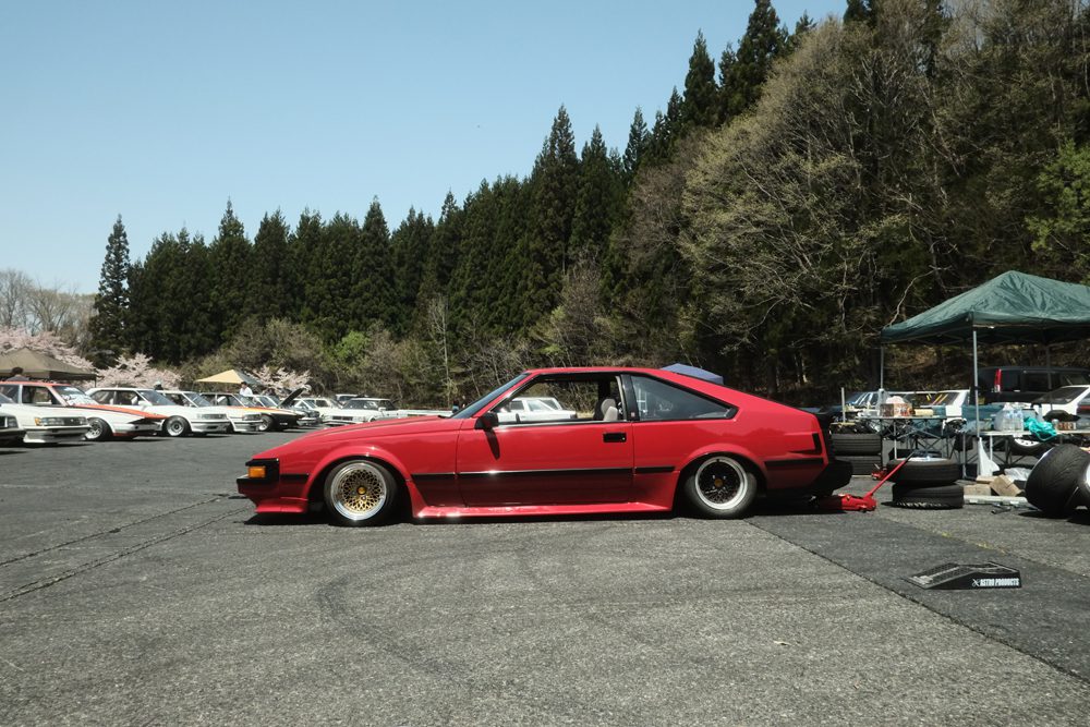 Red classic Toyota Supra at outdoor auto event, parked on asphalt with forest background and other vehicles in view.