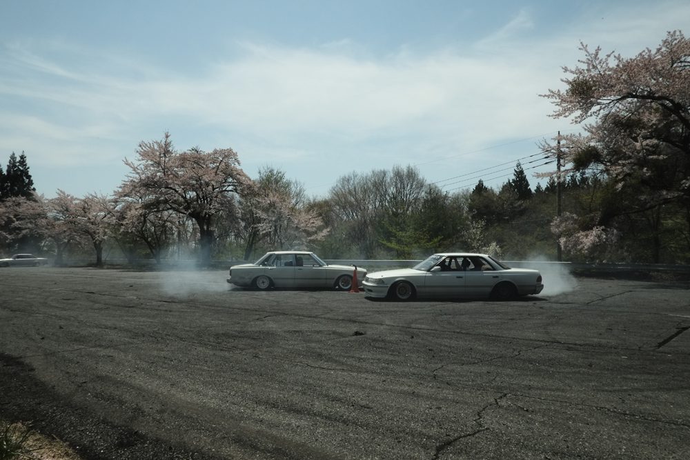 Two cars drifting on an asphalt track surrounded by cherry blossom trees, creating smoke under a clear sky.