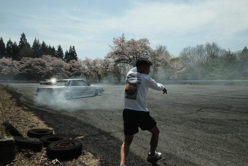 Person observing a car drifting on an open track with cherry blossom trees in the background.