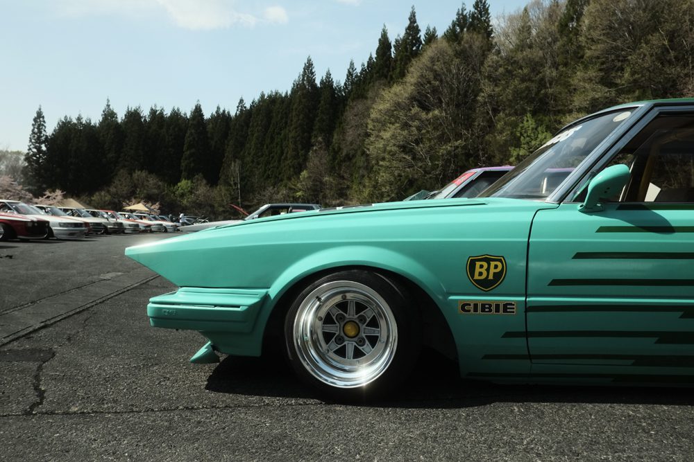 Turquoise custom car with decals at a parking lot, surrounded by trees under a clear sky.