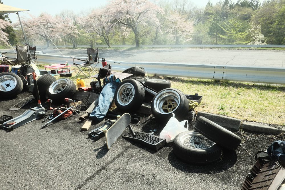 A collection of car tires, tools, and a skateboard on a roadside during a sunny day with blooming trees in the background.