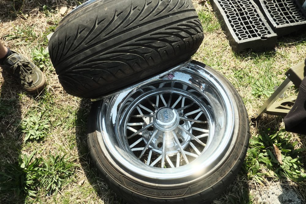 Close-up of a Cragar car wheel with a tire and chrome hubcap on grass, next to shoe and metal structure.