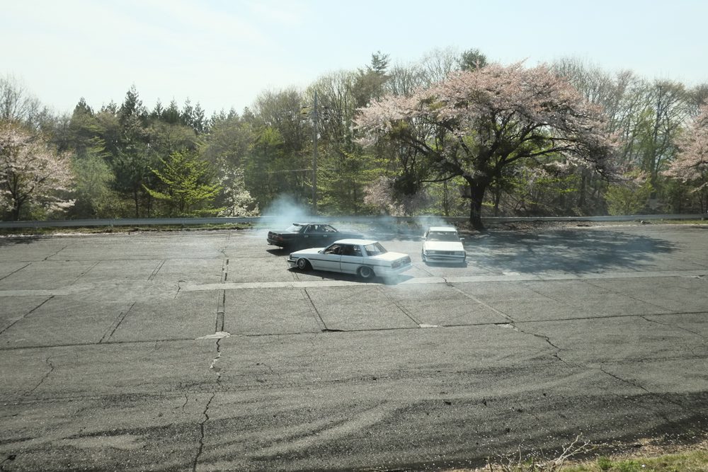 Cars drifting on an open asphalt area surrounded by cherry blossom trees in full bloom.