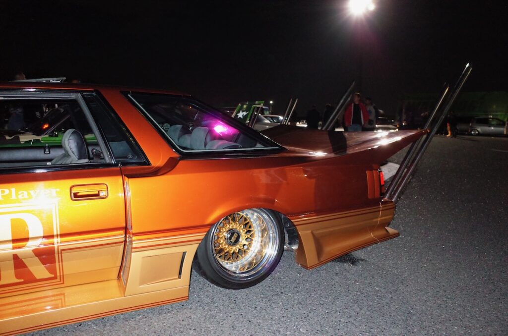 Vintage customized Nissan Leopard with large rear wing and gold rims at night car meet.