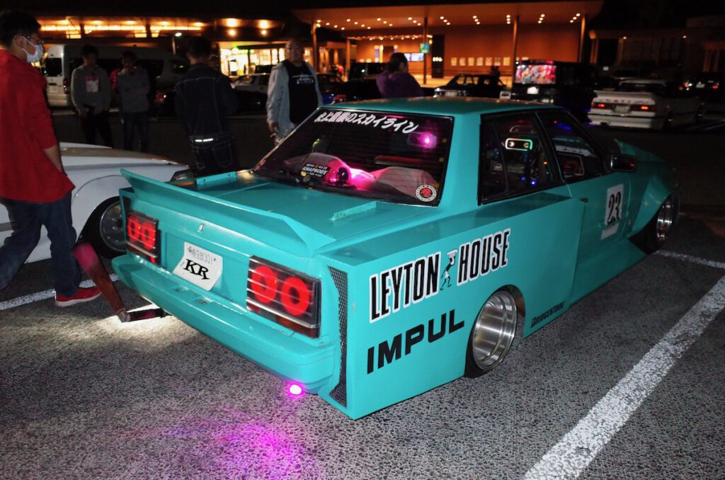 Teal Nissan Skyline racing car with custom decals and neon lights at a night car meet, surrounded by onlookers.