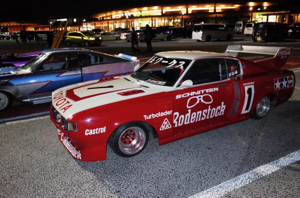 Red and white vintage race car parked at night in a well-lit parking lot.