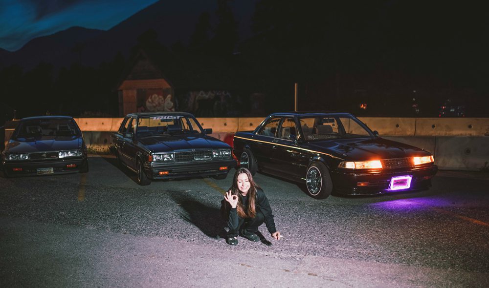 Sandra posing with classic cars at night, illuminated by headlights and purple neon light, in a parking lot.