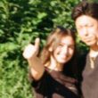 A woman smiling and giving a thumbs-up, standing next to a man with blurred faces in a green outdoor setting.