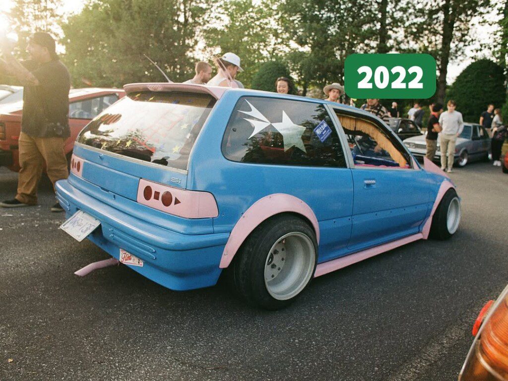 Vintage blue hatchback with pink accents at a 2022 car meet, surrounded by enthusiasts in a park setting.