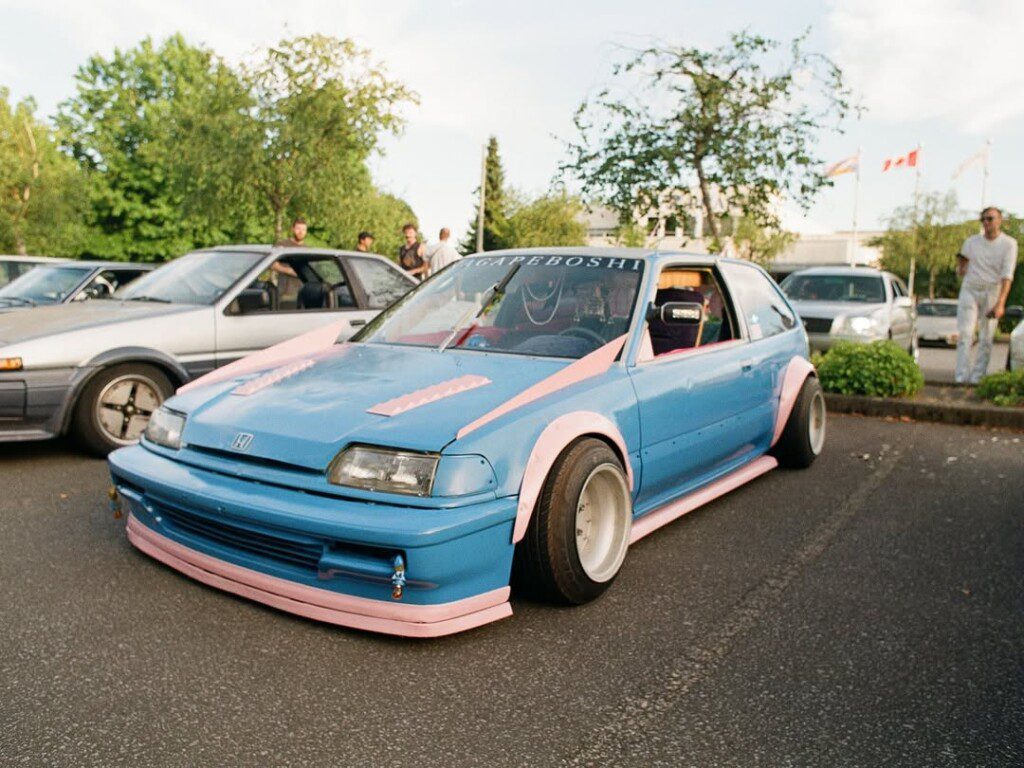 Custom blue and pink Honda hatchback with modified stance at a car meet, surrounded by other vehicles and people.