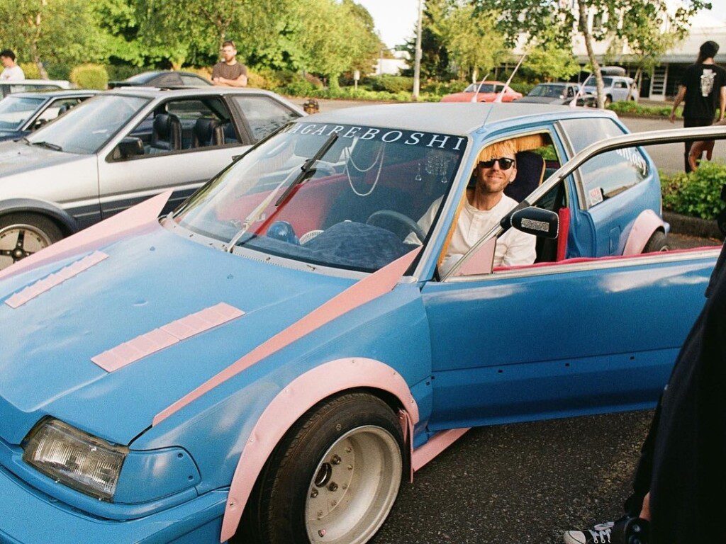 Man smiling from a customized blue and pink car at a car meet with others in the background.