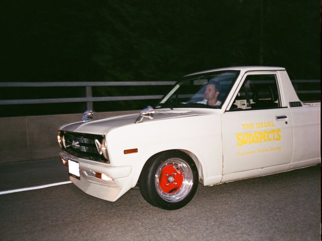 Vintage white pickup truck driving at night, advertising The Usual Suspects on its side with distinctive red wheels.