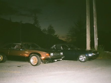 Two vintage cars parked side by side on a dimly lit street at night, surrounded by trees and electricity poles.