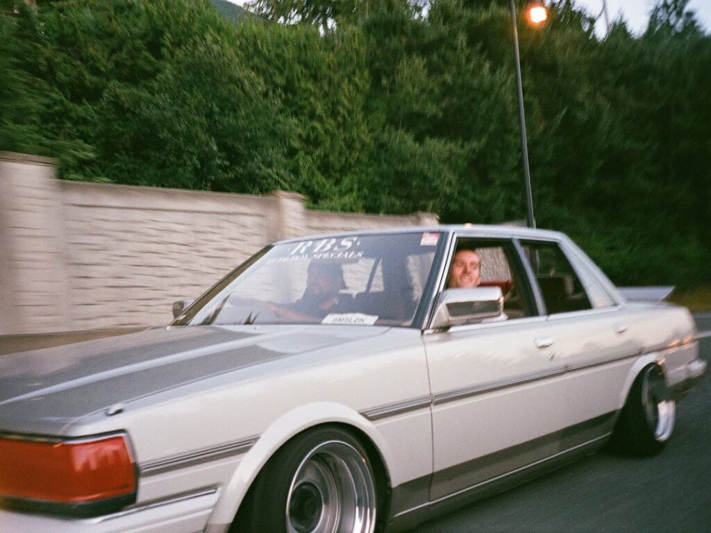 Vintage car cruising on a scenic road with lush green trees and stone walls in the background.