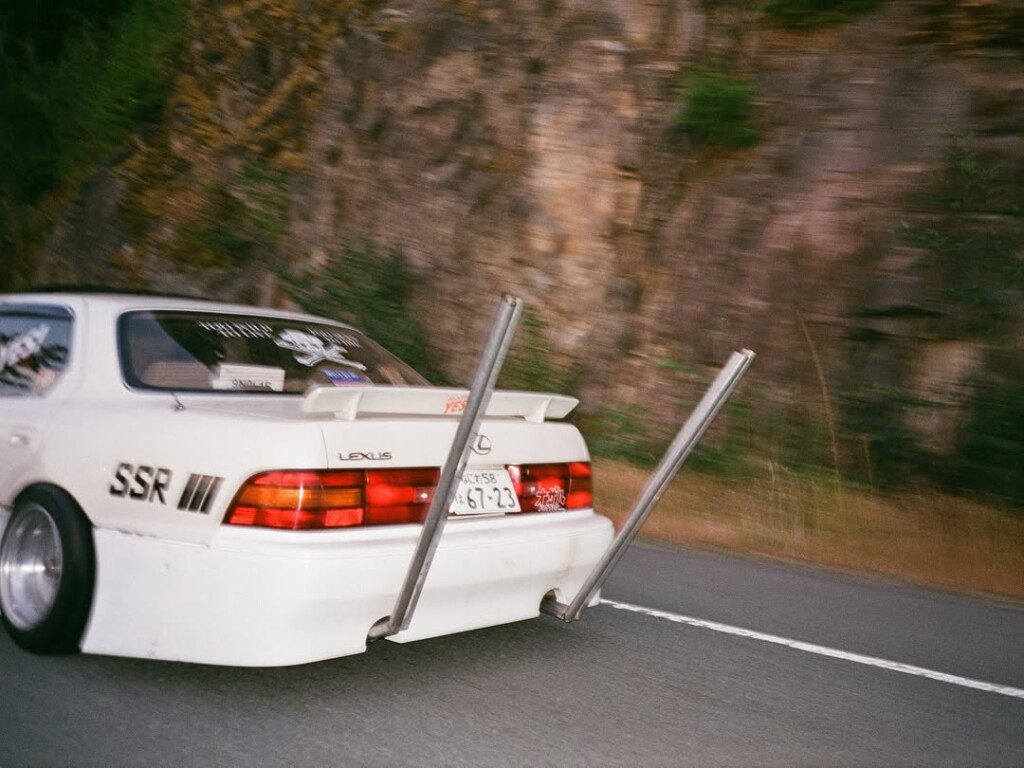 Modified white Lexus car with extended exhaust pipes driving on a highway beside a rocky landscape.