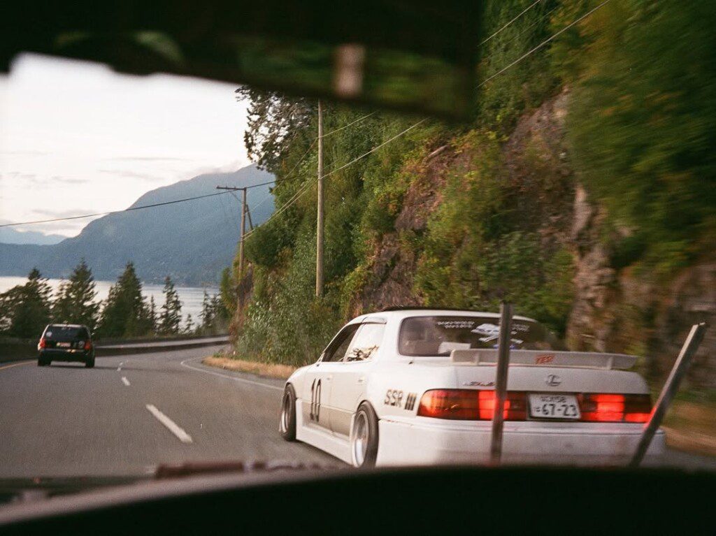 White sports car driving on scenic road with mountain and ocean view, surrounded by lush greenery, under clear sky.