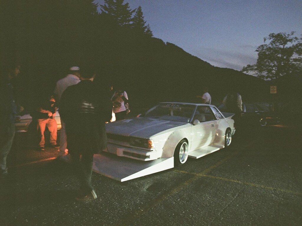Classic car meets at dusk, surrounded by people, under a twilight sky with a wooded mountain backdrop.
