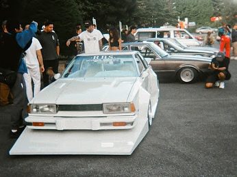Modified classic cars on display at outdoor car show with a crowd of enthusiasts gathered around.