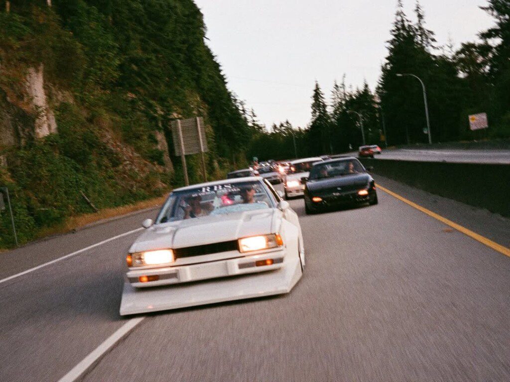Vintage cars cruising on a scenic highway lined with trees, showcasing retro automotive culture.