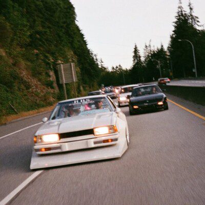 Vintage cars cruising on a scenic highway lined with trees, showcasing retro automotive culture.