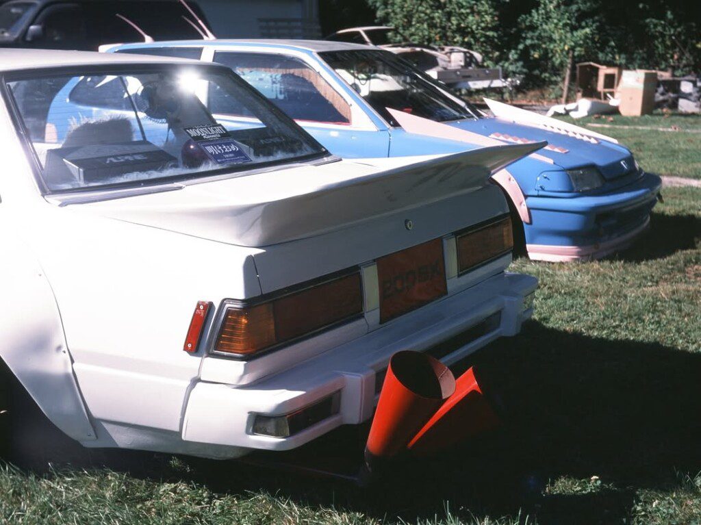 80s modified cars with rear spoilers parked on grass at a car show.
