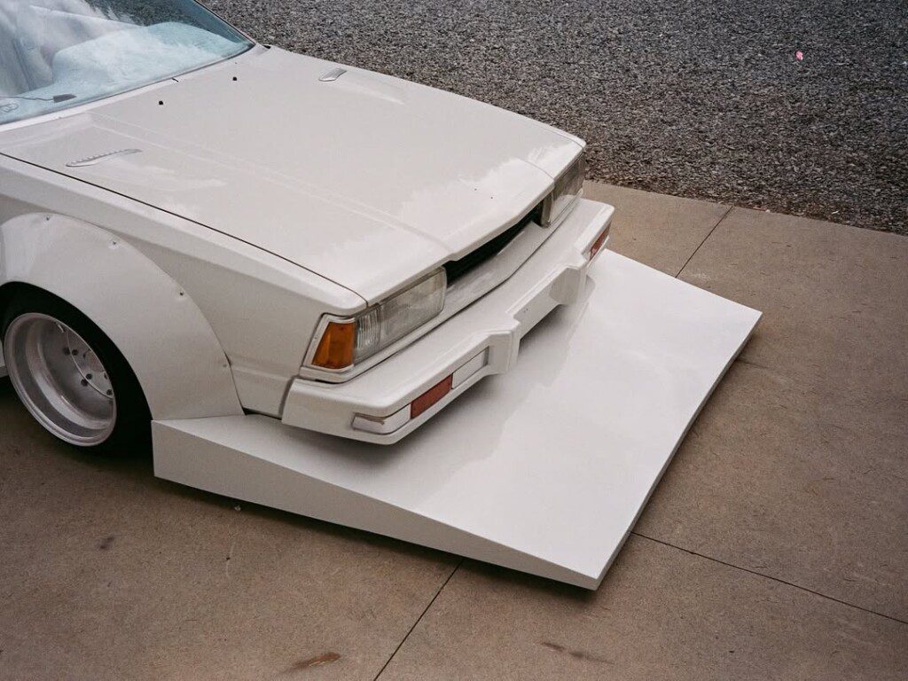 Lowered white car with modified front bumper and flared fenders, parked on pavement, showcasing custom design.