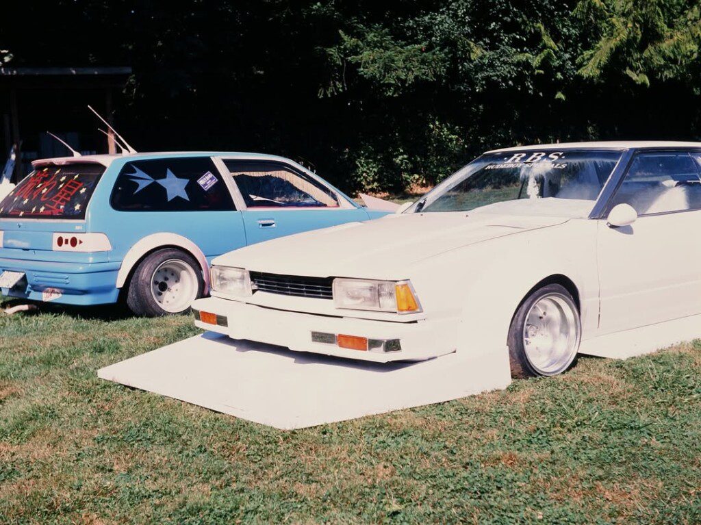 Two modified cars, one blue and one white, parked on grass with creative designs, showcasing custom automotive styling.