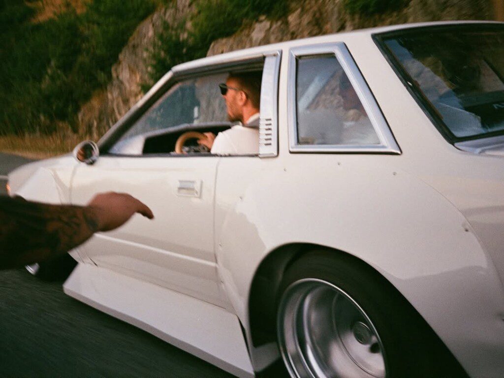 Vintage car driving on scenic road with driver in sunglasses, hand pointing at sleek white vehicle.