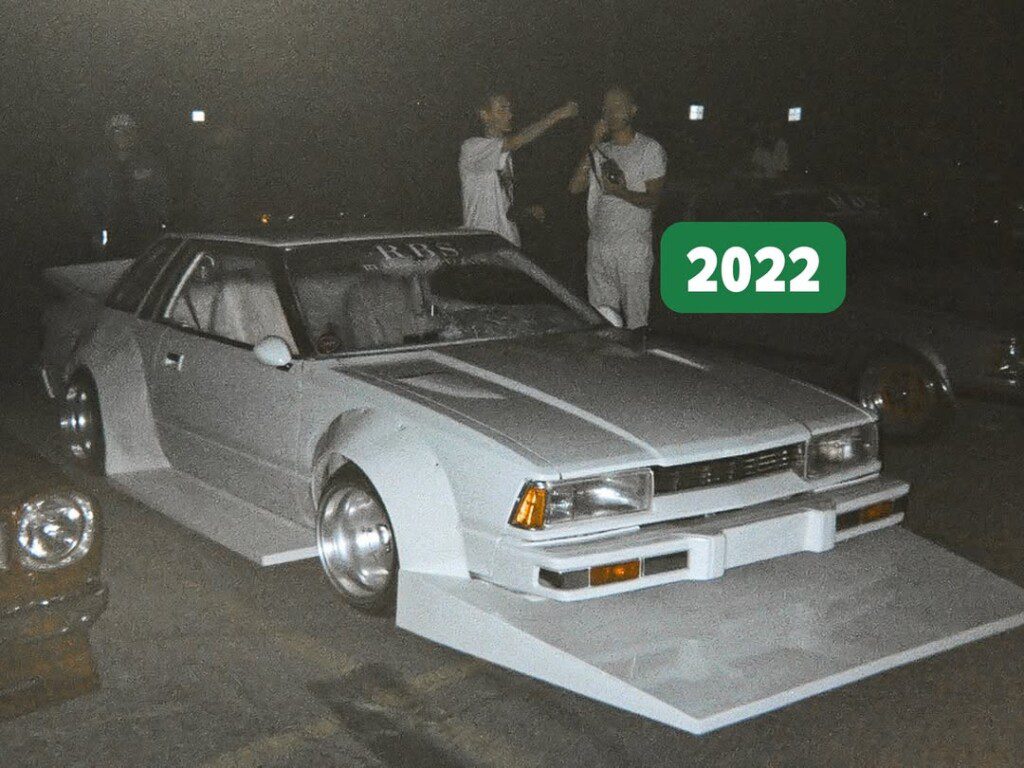 Modified white vintage car with flared body kit at night event, 2022. Car enthusiasts in the background.